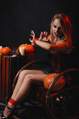 Halloween concept, girl vampire with red eyes red lips sit on rocking chair with pumpkins around. Scary crazy woman trick or treat time. Female makeup for holiday with jake lantern and taped hands