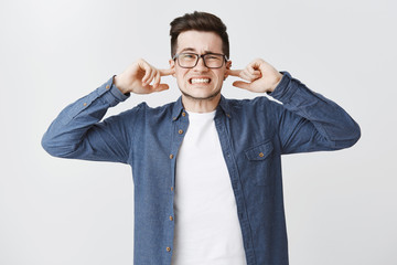 Bothered and irritated upset cute young man in glasses clenching teeth from dissatisfaction frowning closing ears with index fingers being annoyed with loud awful noise over white background