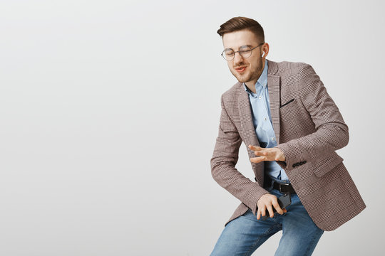 Amused good-looking stylish guy being carried away with cool music bending down in dance move shaking hands to bit of music closing eyes and smiling joyfully listening music in wireless earbuds