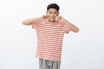 Fototapeta premium Cute chinese guy not used to crowded city life closing ears with index finger not hear loud noise of traffic jams clenching teeth displeased, being distracted with loud awful noise