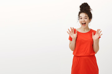 Portrait of surprised thrilled beautiful female with curly hair in casual red dress, raising palms and screaming from positive emotions, being impressed and happy while standing against gray wall