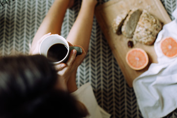 Fototapeta na wymiar Young woman holding a cup of coffee and sitting near a wooden board with sliced freshly baked bread and 2 halves of grapefruit