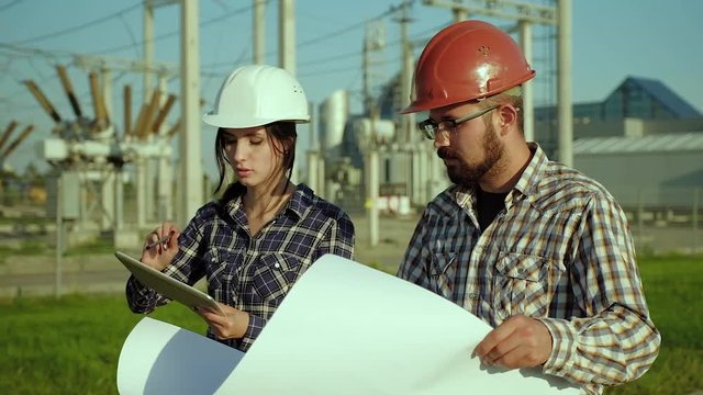 Two engineers discuss on plan on big piece of paper while walking in a field. The plant is on the background. Slow motion