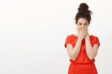 Indoor shot of intense disgusted beautiful urban female in red dress, crossing palms on mouth and...