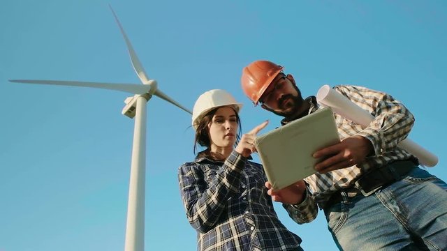 Two engineers in a field are discussing while looking at the tablet. The wind generator is on the background. Slow motion