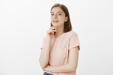 Obraz premium Waist-up shot of feminine confident european female in pink t-shirt, holding hand on chin, smirking and gazing at camera with self-assured expression, being sensual and flirty over gray background
