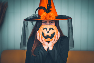 Attractive beautiful Asian woman dressed as a witch holding jack-o-lantern on hand and sitting on yellow sofa, Halloween holidays in cinematic tone. 