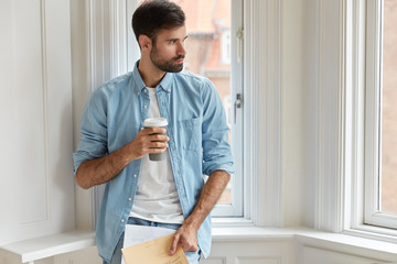 Photo of bearded administrative mamager holds papers, disposable cup of coffee, looks aside thoughtfully, stands near window sill, free space for your advertising content. Entrepreneur does paperwork