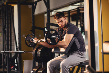 Handsome man doing biceps lifting barbell on bench in a gym