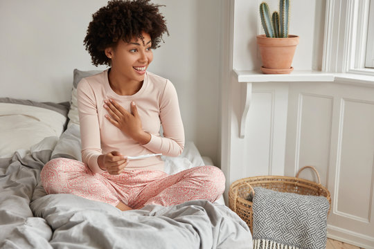 Glaf future mother with Afro hairstyle, dressed in casual pyjamas, sits crossed legs on bed, has satisfied expression, going to share positive news with husband. Feminity and motherhood concept