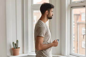 Horizontal shot of young man with dark bristle drinks coffee in morning at modern apartment near window, looks thoughtfully, enjoys domestic atmosphere, deep in thoughts about future career.