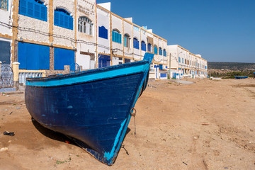 Fototapeta na wymiar A blue boat is stranded on the beach in front of the Tafelney fishing village buildings