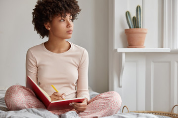 Cropped shot of dark skinned woman sits crossed legs, wears pyjamas, makes notes in diary, focused...