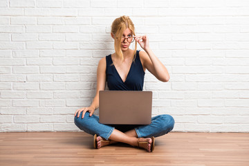 Blonde girl sitting on the floor with her laptop makes funny and crazy face emotion on white brick...