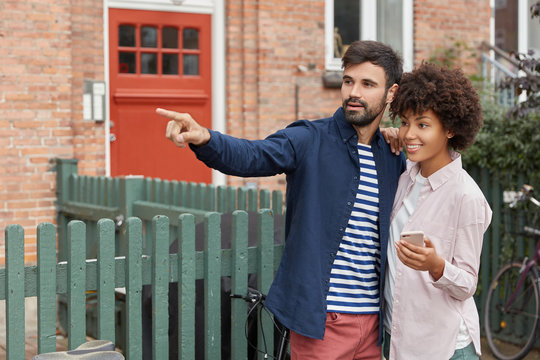 Photo of black woman and Caucasian guy have outdoor walk, stand closely, point into distance as notice friend on other side of road, use smart phone, connected to wireless internet. Livestyle concept