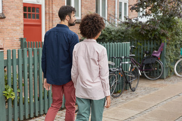 Back view of mixed race couple hold hands, return home after outdoor walk, going to ride bike, wear stylish outfit, have pleasant talk with each other. Relationships, friendship, street style concept