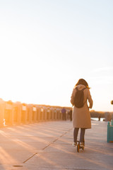 Brunette woman in light beige coat riding scooter in Park in autumn