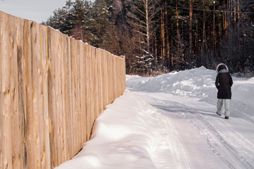 Obraz premium Girl walking on a snowy road along the wooden fence and forest in countryside