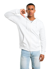 Dark-skinned young man with white sweatshirt showing thumb down sign with negative expression. Sad expression on isolated white background