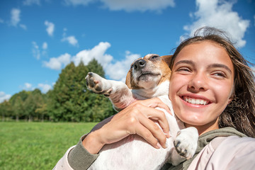 girl hugging her dog and laughs, trees in the park and blue sky background