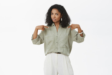 Stylish African-American woman pointing at herself with unsure expression being confused friend blamed het accused in making mistake raising eyebrow curiosly standing doubtful over white background