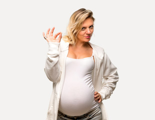Pregnant blonde woman with white sweatshirt showing an ok sign with fingers on isolated grey background