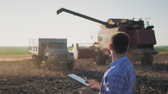 Handsome farmer with tablet standing with combine harvester in background. Farmer use modern technology touch tablet gathering data related to harvest sunflower. Concept the agro-industry.