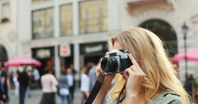 Close up of the pretty smiled Caucasian woman tourist with a blonde hair taking photos on the camera in the town street. Outside.