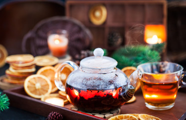 Glass teapot of hot black tea on cozy background with dried oranges and candles