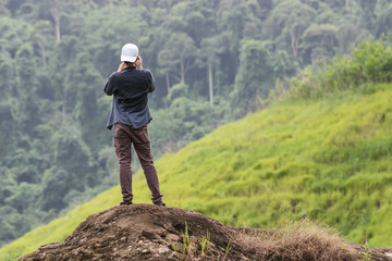 Naklejka premium Back view of young Asian traveler man taking photo on the rock outdoors scenic mountain background. Travel lifestyle and relaxation concept.
