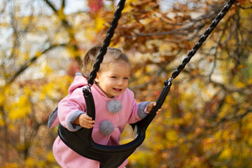 Portrait of a joyful child riding on a swing in autumn park. Smiling child in a pink autumn coat while riding on a swing. Sweet little girl with curls in the wind and smiling eyes.