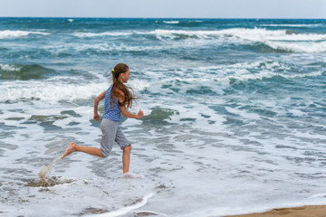 Cute happy little girl running along the beach in jumping over waves. Beautiful summer sunny day, blue sea, picturesque landscape.