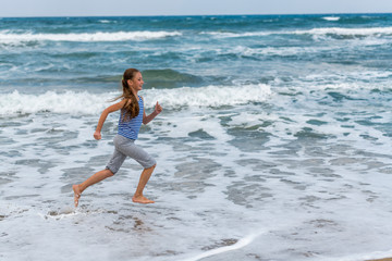Fototapeta na wymiar Cute happy little girl running along the beach in jumping over waves. Beautiful summer sunny day, blue sea, picturesque landscape.