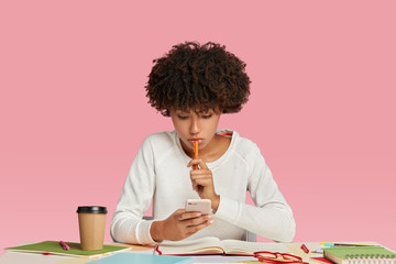 Black female manager looks seriously at smart phone, wears white sweater, keeps pen in hand,...