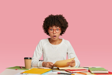 Photo of astonished dark skinned young woman has bated breath, reads unexpected information in paper documents, wears transparent glasses, prepares business project indoor, drinks hot beverage