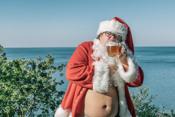 Fat man in glasses dressed as Santa drinking beer on the ocean. Funny, drunk and happy