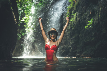 Young woman tourist with straw hat and red swimsuit in the deep jungle with waterfall. Real adventure concept. Bali island.
