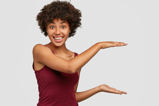 Happy black young woman with pleased facial expression shows height of something, gestures against white background, satisfied with amount, has positive smile on face. Ethnicity and size concept