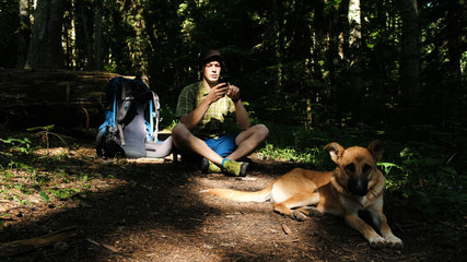 Naklejka na ściany i meble Young man tourist with a dog and a backpack is sitting in the forest and the guy is using a smartphone.