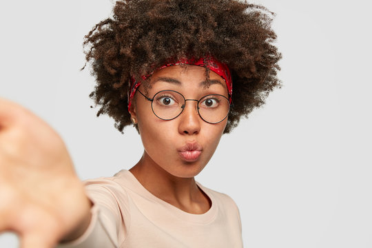 Pretty black oman pouts lips as takes selfie stretches hands in front, wears transparent glasses, casual sweater, isolated over white studio wall. People, ethnicity, facial expressions concept