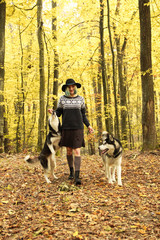 Naklejka na ściany i meble Two Siberian Husky walk in the autumn forest with their mistress. Girl in a hat in a knitted sweater and skirts with leggings. Black and white dogs and feathered leaves.