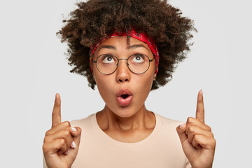 Photo of surprised Afro woman indicates with both index fingers, keeps mouth widely opened, sees...