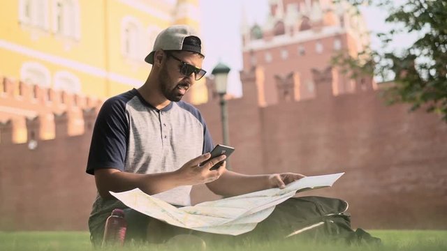 Hispanic tourist wearing sunglasses and baseball cap is looking at his map and using smartphone sitting near Kremlin wall. Handheld slow motion medium shot