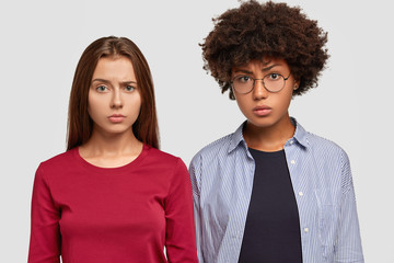Discontent mixed race women frown faces in displeasure, stand closely to each other, recieve bad news, isolated over white background, dressed in casual outfit. People and negative facial expressions