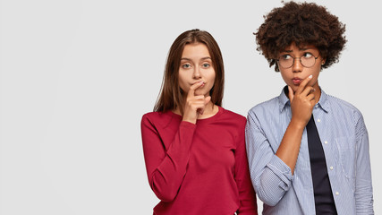 Contemplative two women hold chins, looks thoughtfully, cant make decision, meet to make common task, wear fashionable clothes, isolated over white background with copy space for your promotion