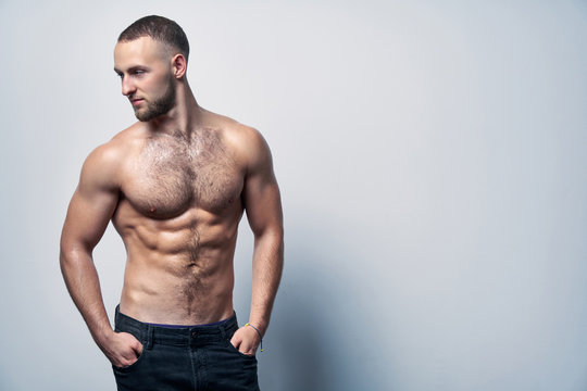 Muscular shirtless man in jeans standing at white wall posing, looking to side