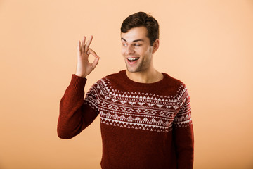 Image of beautiful man 20s with bristle wearing knitted sweater laughing and showing ok sign,...
