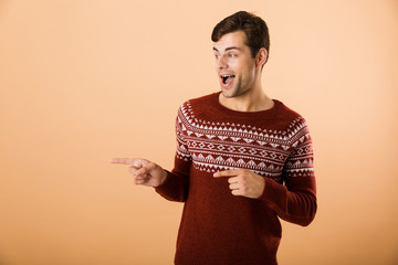 Image of unshaved man 20s with bristle wearing knitted sweater pointing finger aside at copyspace,...
