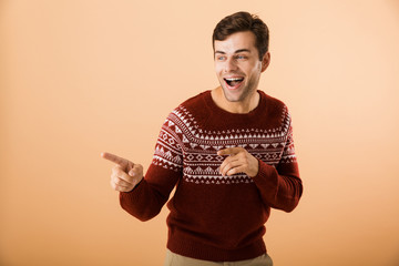 Image of joyous man 20s with bristle wearing knitted sweater pointing finger aside at copyspace, isolated over beige background