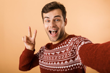 Image of positive man 20s with bristle wearing knitted sweater laughing and showing peace sign while taking selfie photo, isolated over beige background